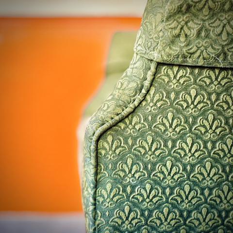 A green, patterned wingback chair is viewed from the left side of its left arm against a short, bright orange wall with white trim above and below.