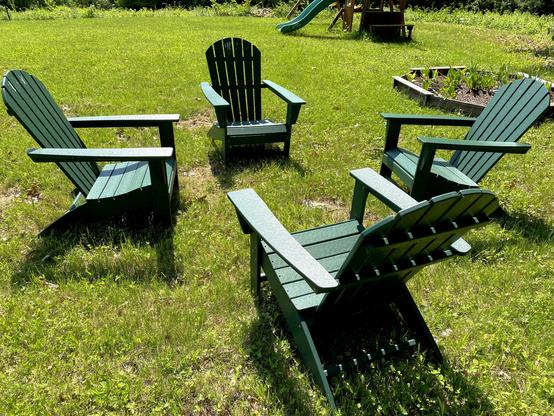Four green Adirondack chairs are arranged in a circular formation on a grassy lawn. In the background, a playground with a slide and a raised garden bed filled with plants can be seen.