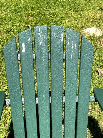 A green Adirondack chair is shown from above, featuring several noticeable marks. The chair is situated on a grassy area.