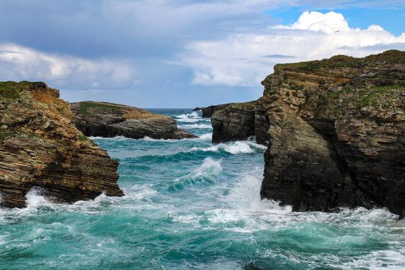 Image of a coastal cliff landscape with waves crashing against them. Above, a blue sky with a few clouds.