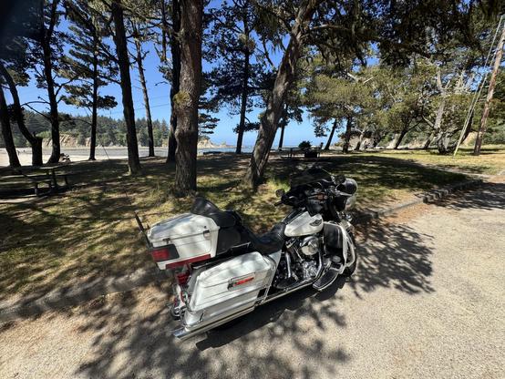 A motorcycle is parked in a shaded area among tall trees, with a beach and ocean visible in the background. The scene features clear skies and nature, suggesting a serene outdoor setting.