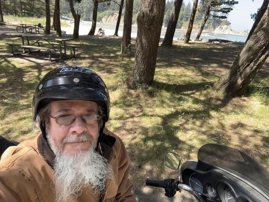 A man with a long beard is wearing a black motorcycle helmet and a brown jacket. He is sitting next to a motorcycle in a shaded area surrounded by trees. In the background, there are picnic tables and a view of a beach or coastline.