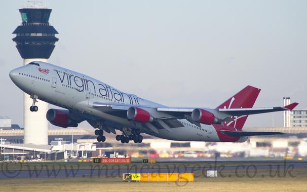 Side view of a very large, 4 engined jet airliner flying from right to left at a very low altitude, with undercarriage extended, flaps deployed from the front and rear of the wings, and the nose raised sharply, suggesting it has just taken off.
The plane is mostly white, with large purple "Virgin Atlantic" titles on the upper forward fuselage, and an image of a flying woman wearing a red leotard and a cape made out of a British flag under the cockpit windows.
The tail and rear fuselage is a vibrant red, with white, hand-written "Virgin" titles.
The engine pods mounted under the wings are the same red, as are the planes up-turned wing-tips.
Green grass fills the foreground, while a grey terminal building stretches across the background, with a tall grey tower with black top floors soars into the grey sky on the left.