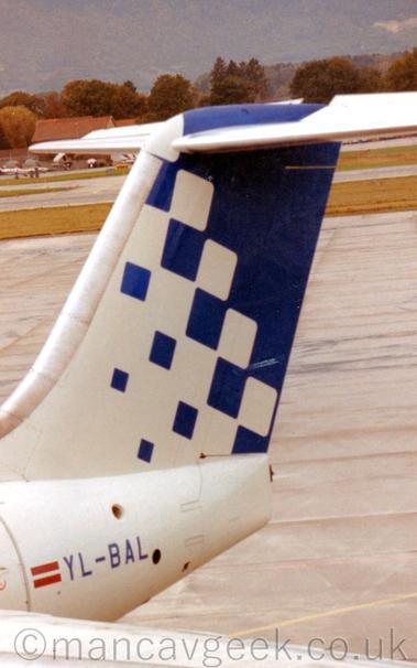 Close-up of the white tail and rear fuselage of a jet airliner parked facing to the left.
Small blue squares at the front of the tail slowly grow bigger as they move rearwards, until they fill the whole of the tail.
The blue registration "YL-BAL" on the rear fuselage, next to a Lithuanian flag.
Grey concrete apron in the background leads off into the distance, with mountains beyond.