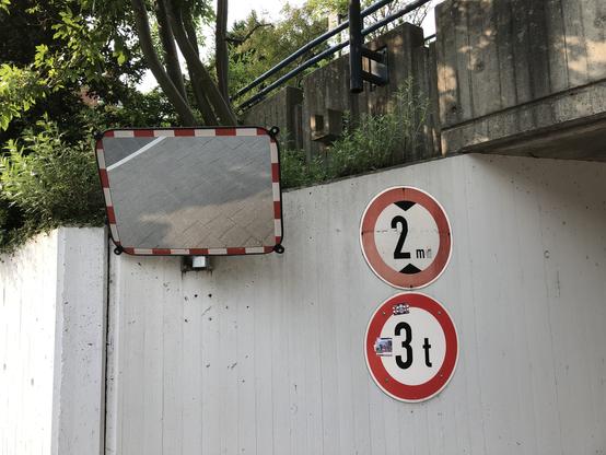 Eine Betonmauer mit großem Spiegel (Seite einer Parkhaus-Einfahrt) mit zwei runder Straßenschildern mit rotem Rand. Das eine zeigt Begrenzung auf 3 t. Oben drüber Grün von Sträuchern.