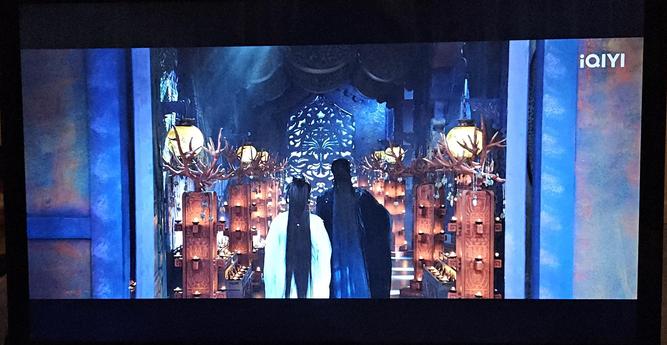 Tien Huo and Fan Yue walk through a room with rust red antlered chandeliers. Round, cream yellow lanterns hang from the blue walls.