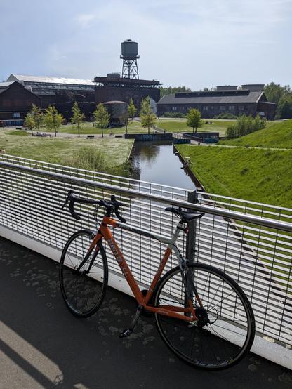 Mein Rennrad steht im Westpark von Bochum an die Brüstung einer Brücke gelehnt.
Im Hintergrund ist die Rückseite der Jahrhunderthalle zu sehen.