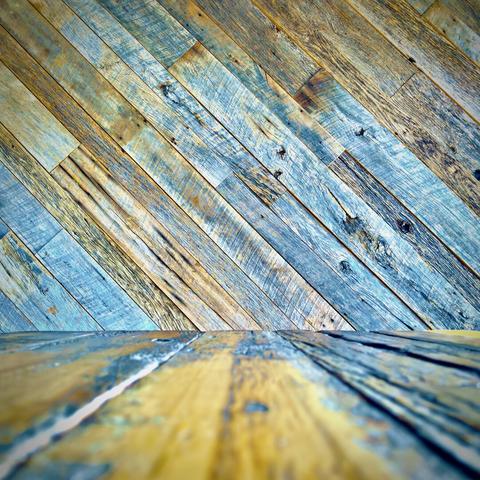 A horizontal wooden floor, comprised of cool-and-warm-tinted two-by-four planks, extends off into the distance, meeting up with a perpendicular wall ahead, comprised of diagonally fitted planks.
