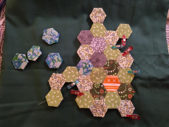 Hex quilt map on a dark green piece of fabric, illuminated in warm artificial light. There's three different textures of green, an orange hex representing rough terrain, the grey forest textures from my last post, and a blue and grey marbled water hex, with more unsewed hexes of the same fabric next to them. A bunch of small clips keep the fabric on the paper in some places.