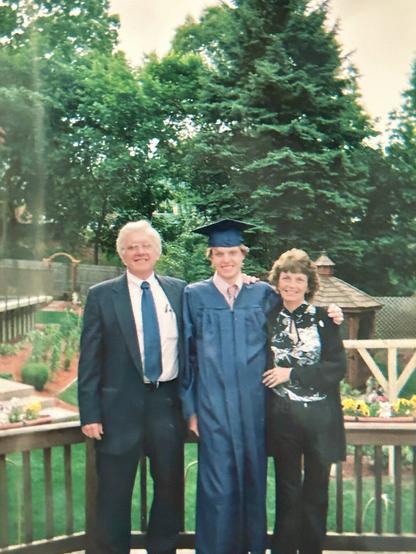 I'm standing in a graduation robe and cap with my father on the left in a suit and tie and my mom on the right in a black blouse with a white design on the front. We are standing on a deck with our backyard behind us in full spring bloom as well as an above ground pool in the back left.