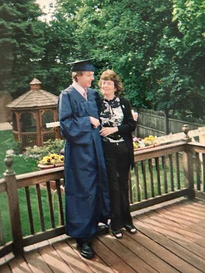 I'm standing in a graduation robe and cap with my mother in a black blouse with a white design on the front. We are standing on a deck with our backyard behind us in full spring bloom. Sunflowers blossom in planters along the railing and a gazebo is in the background.