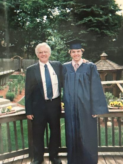 I'm standing in a graduation robe and cap with my father on the left of me in a suit and tie. We are standing on a deck with our backyard behind us in full spring bloom as well as an above ground pool in the back left. Sunflowers blossom in planters along the railing and a gazebo is in the background.