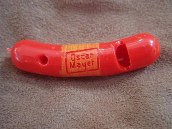 Image of a small, red,  plastic whistle, shaped like a curved hot dog. The "Oscar Meyer" logo is painted/printed in the middle. These came as "prizes" in 8-packs of Oscar Meyer hot dogs in the 1960s, in the USA.