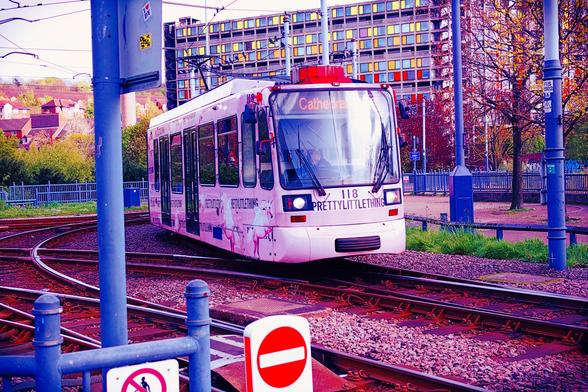Certainly.

A white tram branded "PRETTYLITTLETHING" travels along curved tracks in an urban setting. Its front display reads "Cathedral," indicating its route. The tram is decorated with a stylized unicorn design and moves toward the viewer. Surrounding the tram are overhead wires, metal poles, and a fenced pathway. A large concrete building with colorful window panels dominates the background, contrasting with leafy trees and red-roofed houses further back. The image has a bold, surreal color treatment — purples, magentas, and reds are heavily emphasized, giving the scene a dreamlike, high-fashion atmosphere. A red "no entry" sign appears prominently in the foreground.