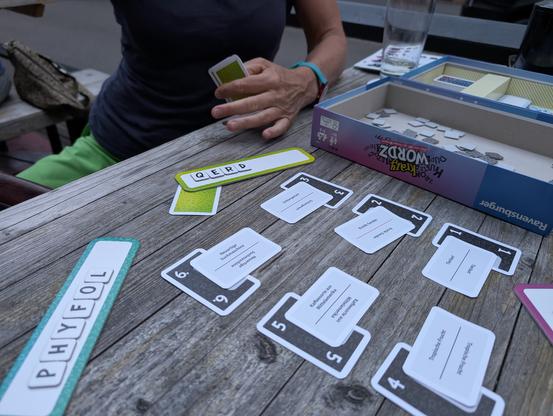 Das Bild zeigt eine Person, die an einem Brettspiel namens "Krazy Wordz" spielt. Die Person sitzt an einem Holztisch und hält eine Karte in der Hand. Auf dem Tisch liegen mehrere Karten und Spielteile des Spiels ausgebreitet. Eine Karte mit dem Wort "QERD" ist sichtbar, sowie eine mit "PHYFOL". Die Karten haben verschiedene Zahlen und Wörter auf ihnen, und einige haben eine grüne oder blaue Umrandung. Im Hintergrund ist ein Spielbox mit dem Namen "Krazy Wordz" zu sehen. Ein Glas Wasser steht auf dem Tisch, und eine Person sitzt am Tisch, die teilweise sichtbar ist. Die Szene wirkt entspannt und spielerisch.