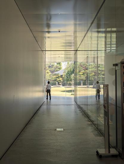 A man walking down a white modern-looking corridor. At the end of the corridor there is a glass wall. Past it a garden with benches and trees can be seen,