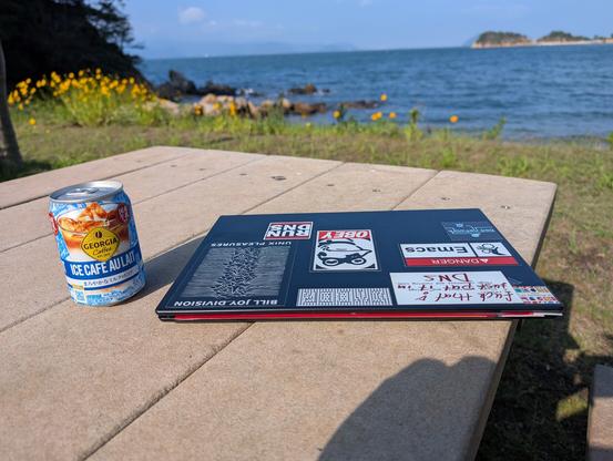 A thin, black Thinkpad laptop with many stickers sits on a stone table at a beach. Blue ocean, blue sky and tiny islands are visible in the background