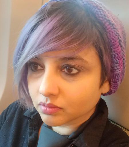 Color close-up of Suzan Palumbo, a Trini woman with dark hair worn in a short asymmetrical bob and tipped with purple. She wears heavy black eyeliner, a black shirt with a collar, a dark gray necktie, and a knit beanie in shades of purple. She looks to the side with a serious expression.
