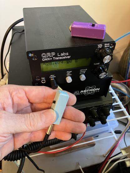 A simple black QMX+ transceiver sits on top of an Astron power supply. It has two rotary knobs on the right, two small gray buttons in the middle for menu navigation, and two 3.5 mm jacks on the left. Above the jacks and buttons is two line LCD screen. Above that is the QRP Labs logo and QMX+ Transceiver name. On top of the QMX+ is a 1.5 inch long microphone with a purple 3d printed assembly. A white guy's hand holds a stone-flecked turquoise color version of the microphone.