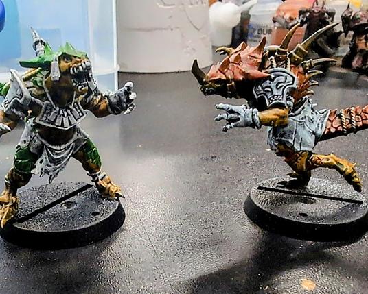 Two Saurus Blocker miniatures from Blood Bowl by Games Workshop. The miniatures have been primed in black and heavily drybrushed with off-white to get ready for "slapchop" painting. The Saurus on the right has yellow skin with orange scales, while the one on the left has yellow skin with green scales.