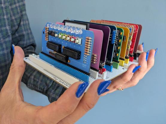 An assembled RC2014 Zed Pro modular Z80 hobby computer, which comprises a backplane about the size of a paperback book with playing-card sized component circuit boards plugged into it vertically. This particular Zed Pro is the limited-edition Pride kit, where the component boards are coloured so that they form a rainbow when arranged in the backplane.

The computer rests in the hands that assembled it, which here are sporting some fairly long, blue-varnished fingernails. All of the electrical components, including dozens of resistors, headers, and IC sockets, were placed and soldered with the fingers and fingernails shown.