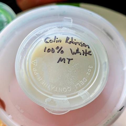 A 4oz condiment cup filled with settled titanium white acrylic paint. The lid has "Colin Robinson, 100% White" written on it in black Sharpie.