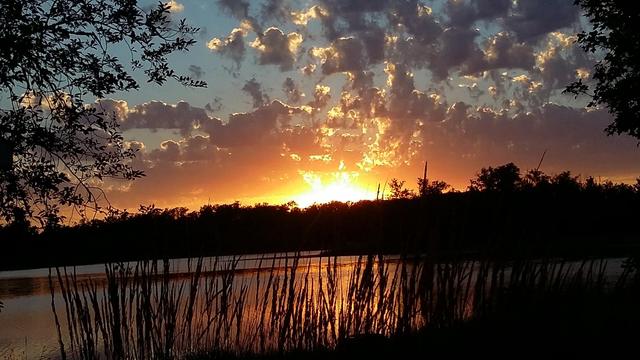 A glowing sun sets over a lake, reflecting a beautiful off of clouds.