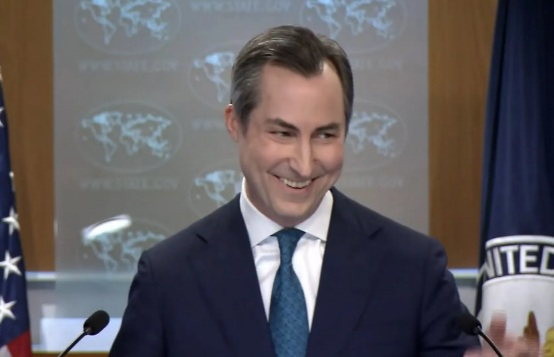 Photo of Matthew Miller at the WH press podium. He is looking to his left and is smiling widely.