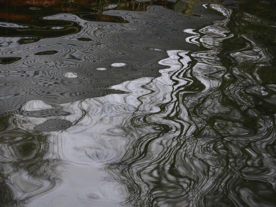 Rippling water surface with distorted reflections of trees and sky, creating intricate patterns. Dark and light areas contrast, emphasizing the swirling, wavy lines across the image.
