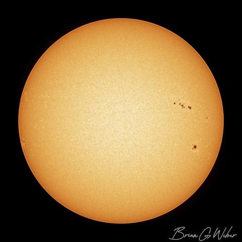 A full disk, white light image of the sun with a line of sunspots above the equator and a larger sunspot below on the right side.