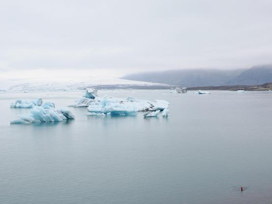 A landscape featuring floating icebergs in a calm body of water, surrounded by brown mountains under a foggy sky. A white glacier is visible in the background. A lone swimmer in a red wollen hat is visible in the water.