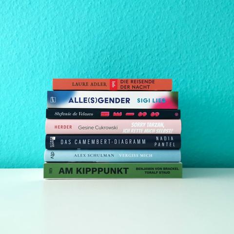 Auf dem Foto ist ein Bücherstapel aus sieben Büchern vor einer türkisen Wand auf einer glatten, weißen Oberfläche zu sehen. In der Caption werden die Bücher näher beschrieben.