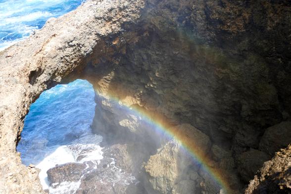 Der Betrachter steht oben am Rand einer schmalen Felsspalte, die sich zum Ozean hin öffnet. Ein Felsbogen überspannt diese Öffnung. Die brechenden Wellen erzeugen einen Regenbogen innerhalb der Felsspalte.