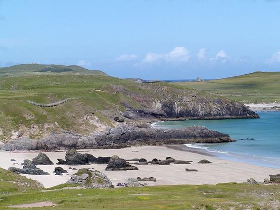 Sango Bay at Durness. The image shows a view over a sandy beach with dark rocks in it to a series of grassy headlands with dark rock edges. The sea is to the right of the frame and is turquoise. The scene is in sunlight.