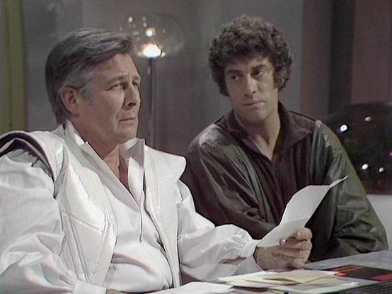 Claude 3.7 describes the image as: "This image appears to be from a classic British science fiction television series from the late 1970s/early 1980s. The scene shows two men in what looks like a futuristic or institutional setting. On the left is an older man wearing a white uniform or outfit, possibly representing authority or medical profession, holding what appears to be a document or paper. To his right is a younger man with curly dark hair wearing a dark shirt or jacket, who seems to be looking at the document with concern or interest. The lighting and film quality have the characteristic look of BBC productions from that era. The setting suggests this might be taking place in some kind of administrative or control room of a spacecraft or space station."

Blakes7Bot, version 3.9.30.