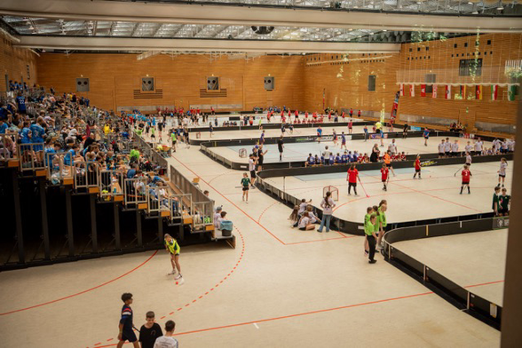 In einer sehr großen Sporthalle ist eine Tribüne mit vielen Menschen und Doktor davor stehen mehrere Floorball Spielfelder, auf denen gerade gespielt wird.
