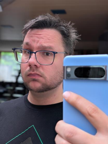 Man with short dark hair, some grey, wearing rectangular glasses, looks at the camera while taking a selfie. He holds a light blue smartphone, and the early growth of a beard is visible on his face. He is wearing a dark t-shirt, and the background is softly blurred.