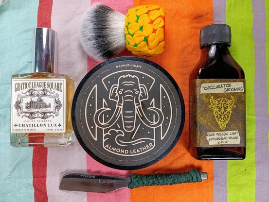 (Le Chat Mistral description)

Thank you for the clarification. Here's the updated description:

The image showcases a collection of grooming products arranged on a striped cloth. At the center, there's a round tin of almond leather-scented soap with a mammoth illustration. To the left, there's a bottle of Gratiot League Square Eau de Toilette by Chatillon Lux. Above the soap, there's a shaving brush with a yellow handle. To the right, there's a small bottle labeled "Declaration Grooming" with a bison head logo, containing fake yellow light aftershave splash. Below the soap, there's a traditional Japanese kamisori with a green handle. The items are neatly organized, suggesting a traditional shaving setup.