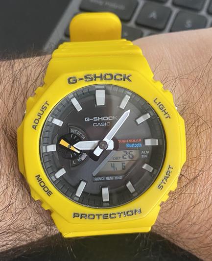 Un reloj gshock amarillo de forma octogonal. Se puede ver el desgaste por el uso en varias de las serigrafías de la caja. El dial es negro y las agujas marcan aproximadamente las 9:05. Abajo a la derecha hay una pequeña pantalla digital con el segundero y la fecha.