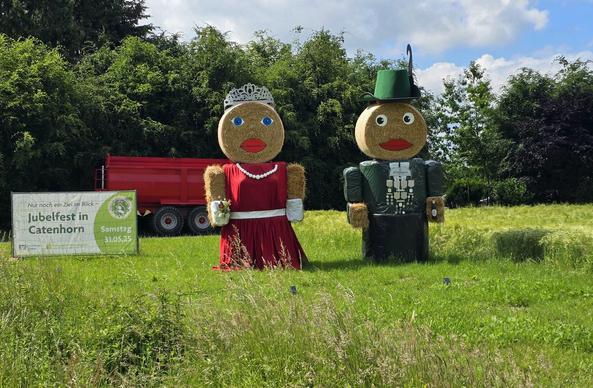 Zwei große Figuren aus Heuballen stehen auf einem grünen Feld. Die linke Figur ist als Königin mit rotem Kleid, weißer Schärpe und einer Krone verziert. Die rechte Figur ist als König mit grünem Hut und traditioneller grüner Tracht dargestellt. Im Hintergrund ist ein roter Anhänger und ein Schild zu sehen, das auf ein "Jubelfest in Catenhorn am Samstag, 31.05.25" hinweist.