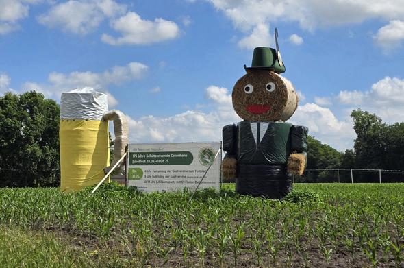 Auf einem Feld mit jungen Pflanzen stehen eine große Heufigur und ein riesiger Bierkrug aus Strohballen. Die Heufigur trägt einen grünen Hut mit Feder und einen schwarzen Anzug mit weißem Hemd. Rechts von den Figuren steht ein weißes Plakat, das auf das "125 Jahre Schützenverein Catenhorn Jubelfest 28.05.-01.06.25" hinweist. Der Himmel ist blau mit weißen Wolken.
