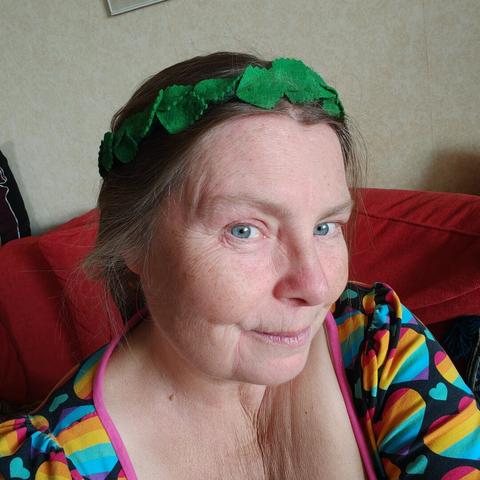 A white middle aged woman is wearing a wreath of green wool fabric leaves. She is smiling slightly.