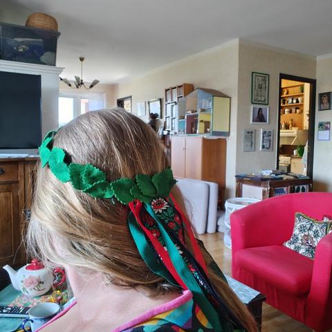 The back of the head of a white middle aged woman. She is wearing a wreath made from various textiles. The wreath is covered in bright green birch type leaves, and has ribbons hanging from it in the back. Red shiny ribbons, green cotton tape, and a jaquard ribbon with red and white flowers, and green leaves, on a black background.