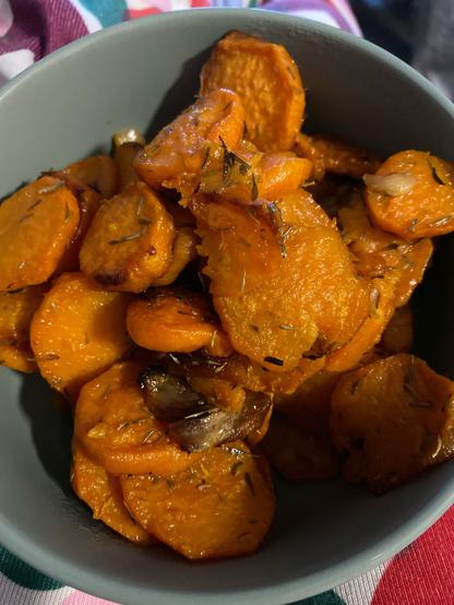 A bowl full of roasted sweet potato slices. They’re glossy and slightly caramelized, dotted with thyme, and there are a few crushed garlic cloves visible.