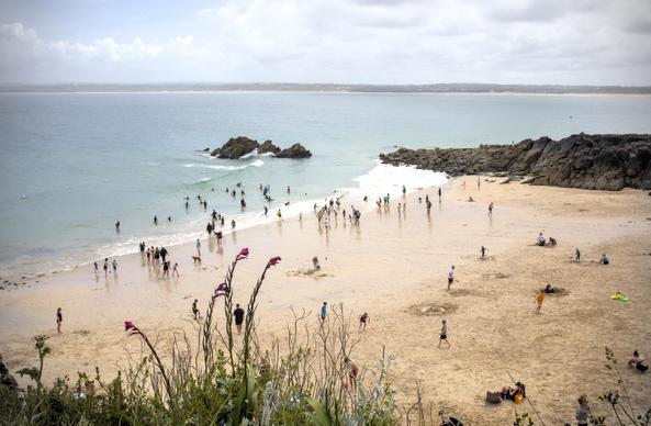 A small beach, taken from a cliff above. People are on the beach and in the sea, playing beach games and body boarding. There are rocks the other side of the beach and some jutting up out of the water. In the background, beyond the sea, more land can be seen - perhaps the other side of a bay.