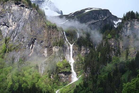 A Y-shaped waterfall falling some 150 m of a limestone cliff. Snow capped mountains in the background and some clouds around the top of the waterfall. Below are deciduous and coniferous trees.