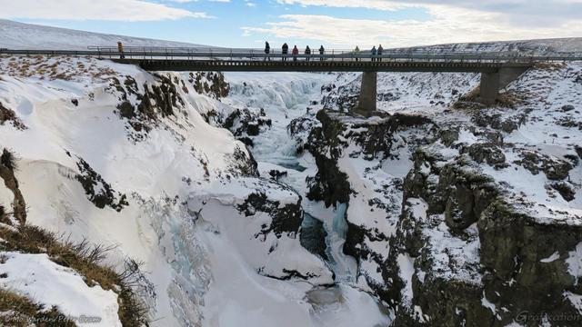 A colour photo of a landscape dominated by a canyon with a bridge across it. The sky is clear but cloudy, and a lovely waterfall in the centre of the shot is frozen almost solid. People are standing on the bridge. The steep walls of the gorge are covered in ice and snow.