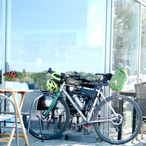 Gravelbike mit Bikepackingausstattung lehnt an einer Glasfase eines Cafes.