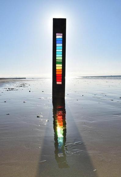 Photo of a slim rectangular wooden sculpture with central stained glass in rainbow colours the artwork is on a beach with wet sand and under a blue sky