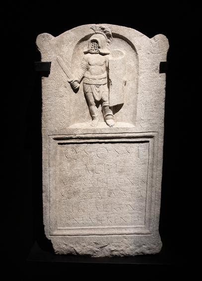 Funerary stele depicts a portrait of a murmillo gladiator at the top followed by the Latin inscription “D (is) M (anibus) / Q (uinti) Sossi / Albi / myrmillonis / Sossia Iusta / lib (erta) patron (o) / meritorious.”  This translates roughly as, “To the Shades of the Underworld, Sossia Iusta freedwoman dedicates this to the worthy patron Quintus Sossius Albius.” Image credit: Dorieo, Wikimedia Commons (License CC-BY-SA 4.0).
